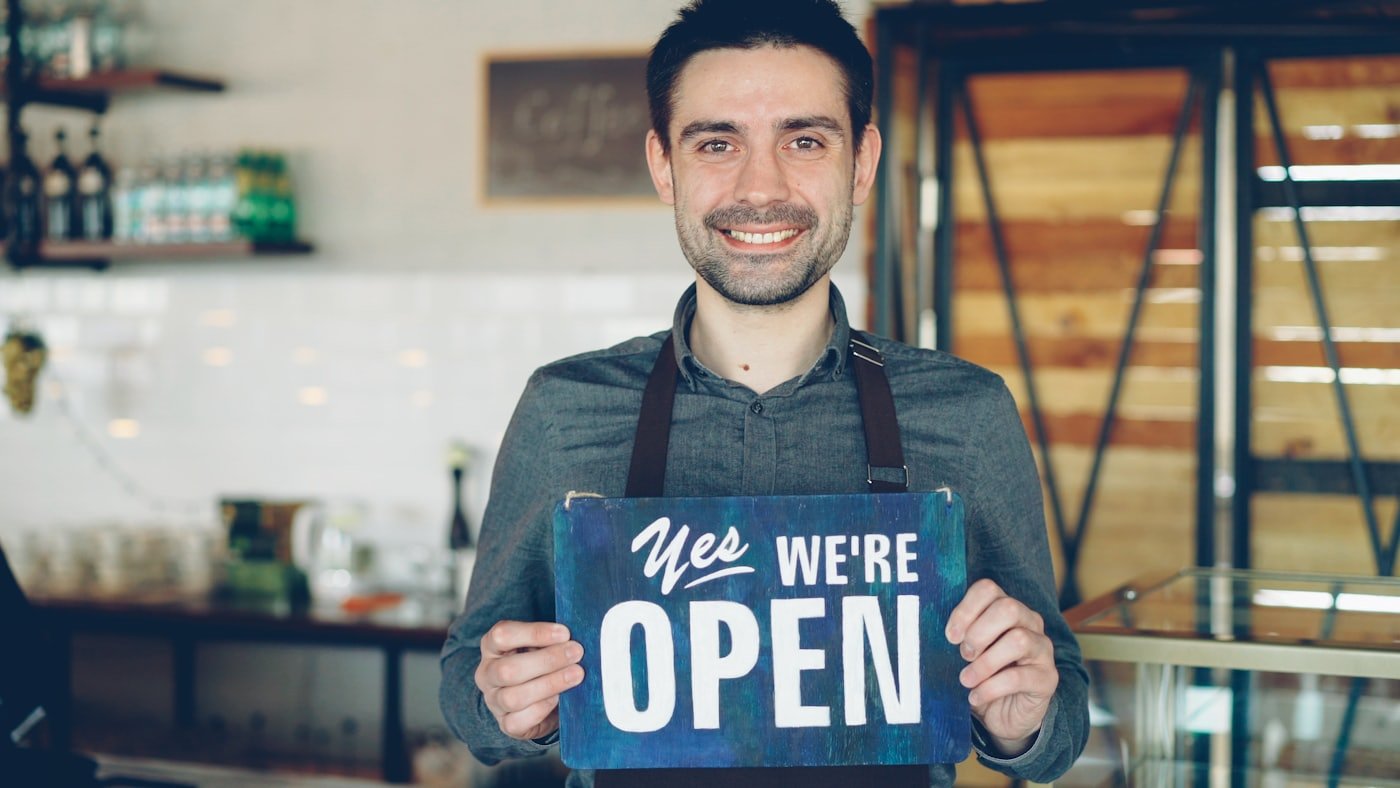 A smiling small-business owner holding an open-for-business sign in their shop